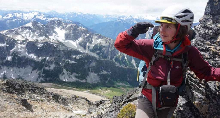 a woman wearing mountaineering gear shades her face with her hand. Snowy mountains are in the background.